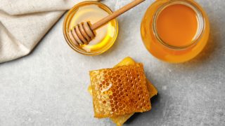 Flatline view of a jar of honey, bowl of honey with honey dipper and a honeycomb on a slate background.