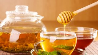 Honey being poured into a cup of tea with mint with kettle and a cup of tea in the background.