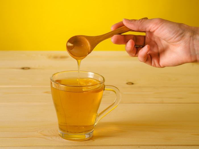 Honey being poured from a wooden spoon into a glass cup of amber liquid containing a citrus slice