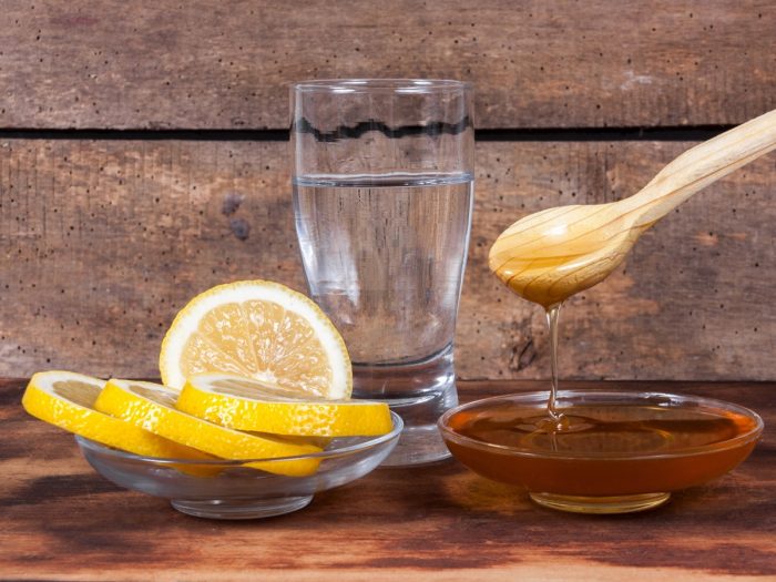 A glass of water, flat bowl of sliced citrus fruits, and a bowl of honey on a rustic background.