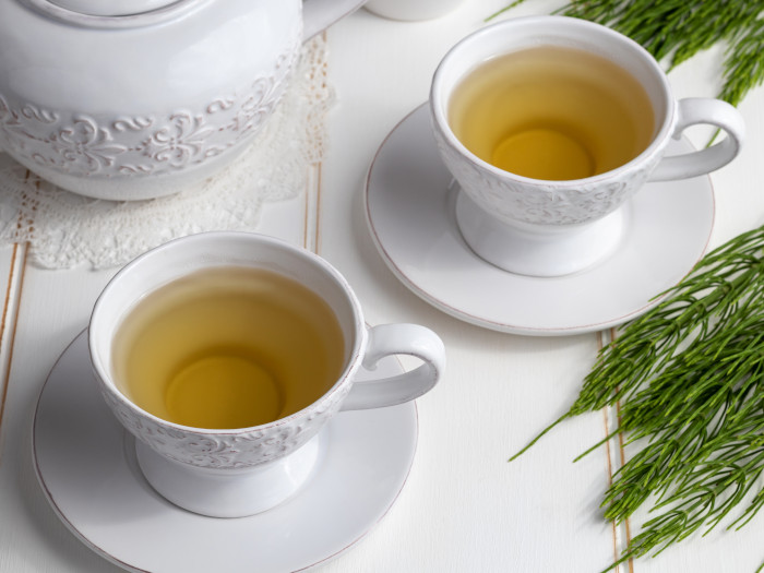 Close-up of two cups and saucers of tea and kettle at the back on a white surface.