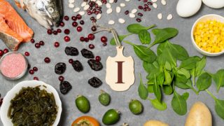 Flatline pic of hand holding a card saying 'I' surrounded by dried fruits, fruits, fish, greens, egg, seaweed, and salt, placed on a blue surface.
