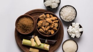 A wooden plate with halved sugarcane and wooden bowls filled with jaggery cubes and jaggery powder, and bowls containing sugar cubes and powdered sugar