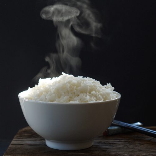 A close up shot of steaming hot cooked jasmine rice in white bowl against a black background.