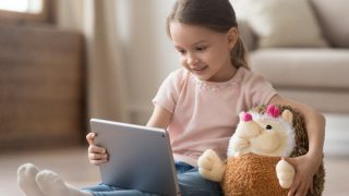 Curious little child girl having fun using a digital tablet and embracing a toy sitting on the floor