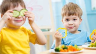 Boy holding cucumber slices on his eyes and another boy holding sliced veggies with plates of food in front of them