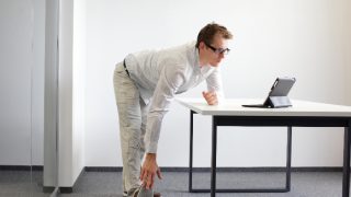 Man standing at a desk looking at a laptop while bending down to touch the toes of his extended right leg