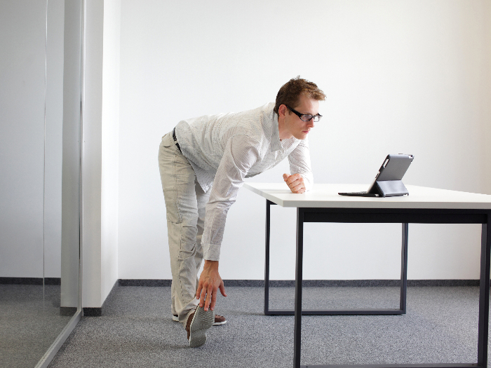 Man standing at a desk looking at a laptop while bending down to touch the toes of his extended right leg