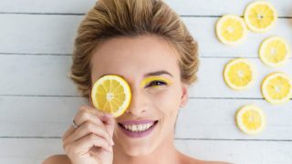A flat lay pic of a smiling woman covering one of her eyes with a lemon slice