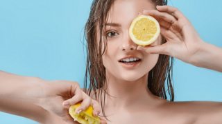 A smiling woman holding a lemon half over an eye and a squeezed lemon in the other hand.