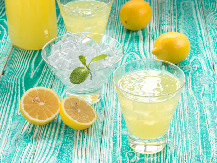 Glasses of limoncello kept besides lemons and a bowl of ice, atop a blue surface