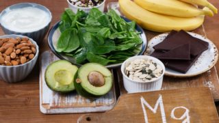 Magnesium-rich foods such as almonds, bananas, legumes, spinach, dark chocolate, and avocado on a wooden table