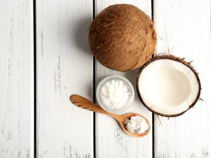 A top view of a small glass jar & a spoon filled with solidified coconut oil, next to 2 coconuts against a white background