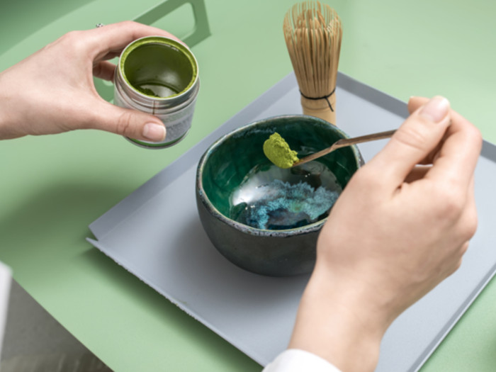 Close-up of someone holding a tin of matcha and a spoonful of matcha over a green ceramic bowl and a whisk placed nearby