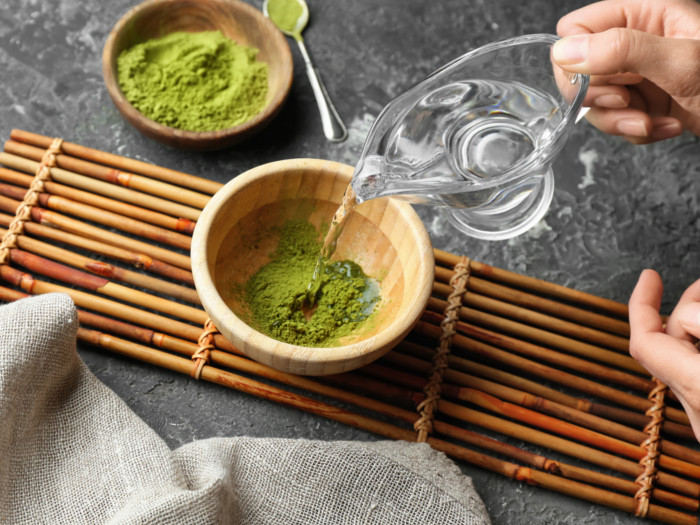 Someone pouring into a bowl of matcha placed on a bamboo mat with a bowl of matcha and spoon at the background.