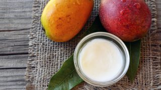 Two mangoes kept next to a bowl of mango butter atop a mat on a wooden table