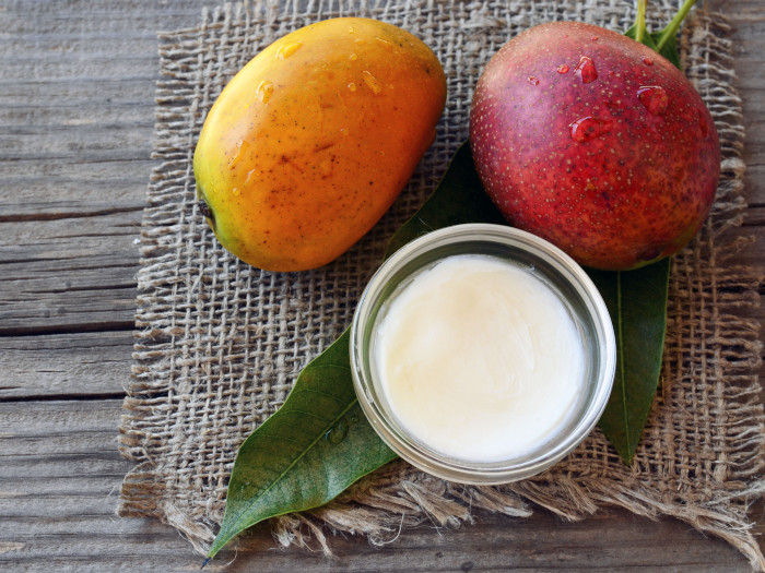 Two mangoes kept next to a bowl of mango butter atop a mat on a wooden table