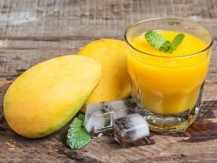 Mango juice in a glass, whole mangoes, and ice cubes on a wooden counter