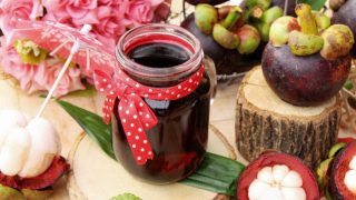 Jar of mangosteen juice placed on a small wooden board, surrounded by mangosteens and a bouquet of flowers at the back.