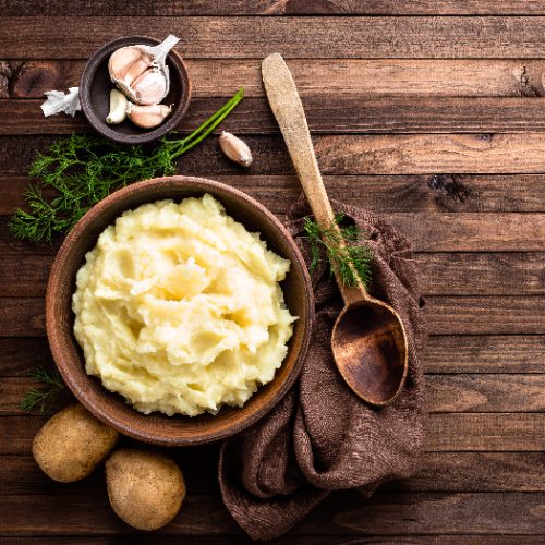 A flatlay picture of a plate of mashed potatoes placed next to two potatoes, brass spoon, napkin, garlic, atop a wooden table