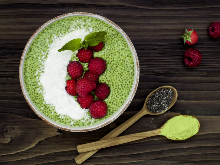 A chia pudding bowl topped with raspberries, more raspberries, spoon of chia seeds and a spoon of yellow powder.
