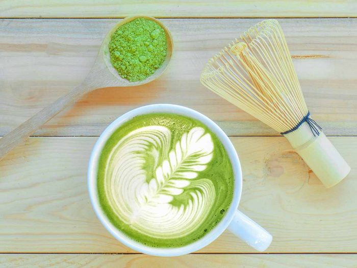 Flatline picture of a spoon of matcha powder, a cup of matcha tea and wooden whisk.