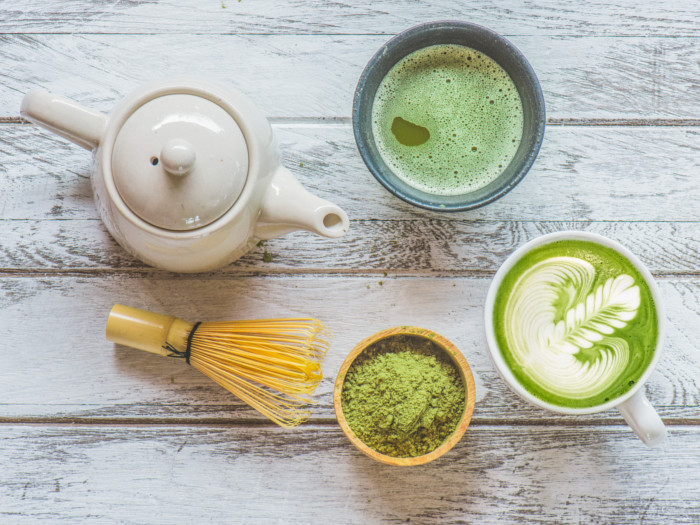 Cups of matcha tea and matcha milk tea with a teapot, brewer, and matcha powder on a wooden table