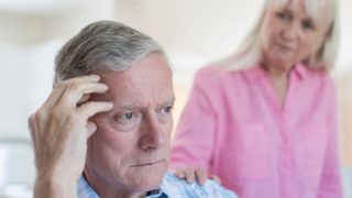 An old man holding his head with a woman at his back