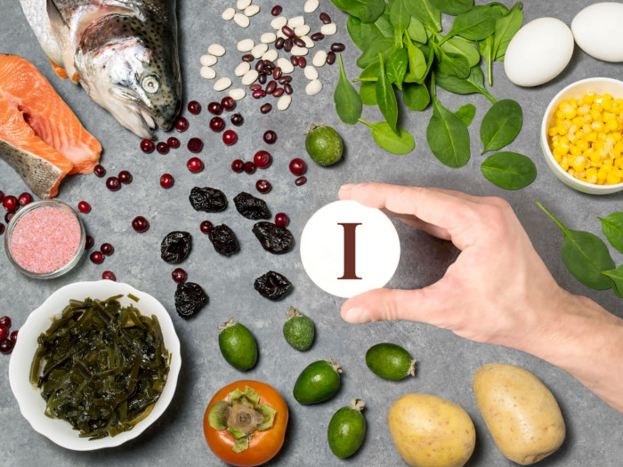 Flatline pic of hand holding a card saying 'I' surrounded by dried fruits, fruits, fish, greens, egg, seaweed, and salt, placed on a blue surface.