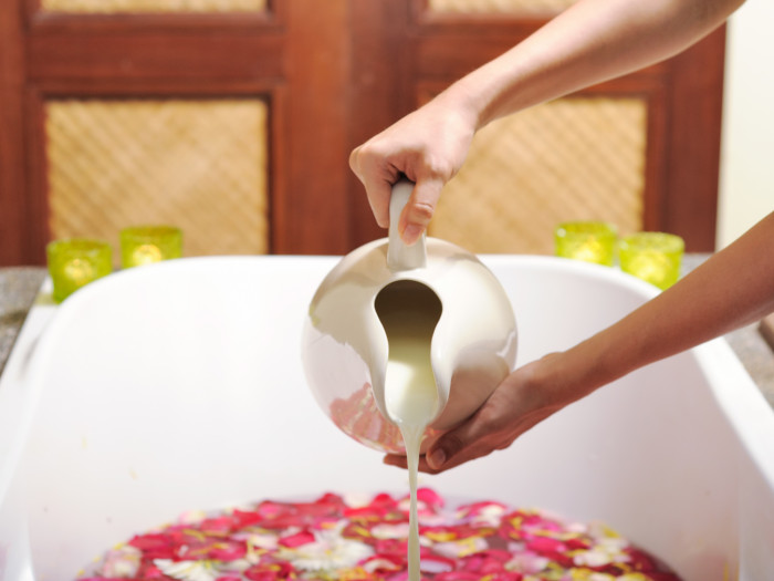 A jug full of milk being poured into a bathtub decorated with rose petals