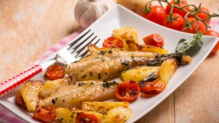 Cooked fish with veggies and potatoes on a plate, served on a wooden surface with tomatoes on the vine, a potato, and garlic.