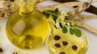 A cup of moringa oil with moringa seeds in it, a jar of moringa oil and flowers on a white background
