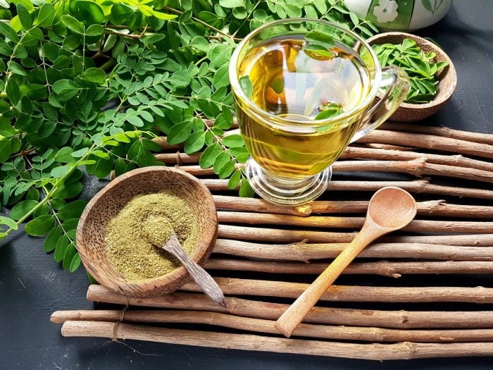 A cup of moringa tea with a wooden bowl of moringa powder and fresh leaves on wood sticks