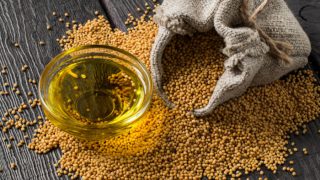 A bowl of mustard oil, yellow mustard seeds in a burlap bag as well as scattered on wood
