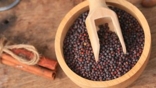 Flatline picture of a small bowl of mustard seeds with a bundle of cinnamon sticks nearby on a wooden surface.