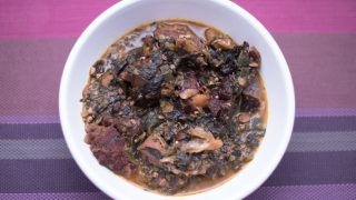 A white plate of Ogbono soup with mixed okra, dried fish and and bush meat on a purple mat