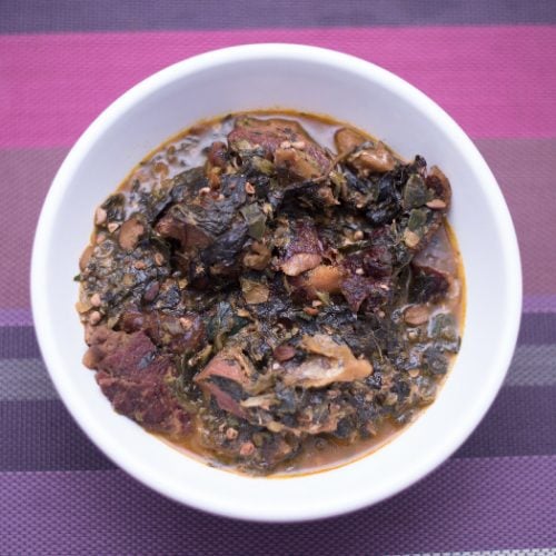 A white plate of Ogbono soup with mixed okra, dried fish and and bush meat on a purple mat