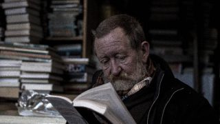 Old man reading a book with a stacks of books in the background