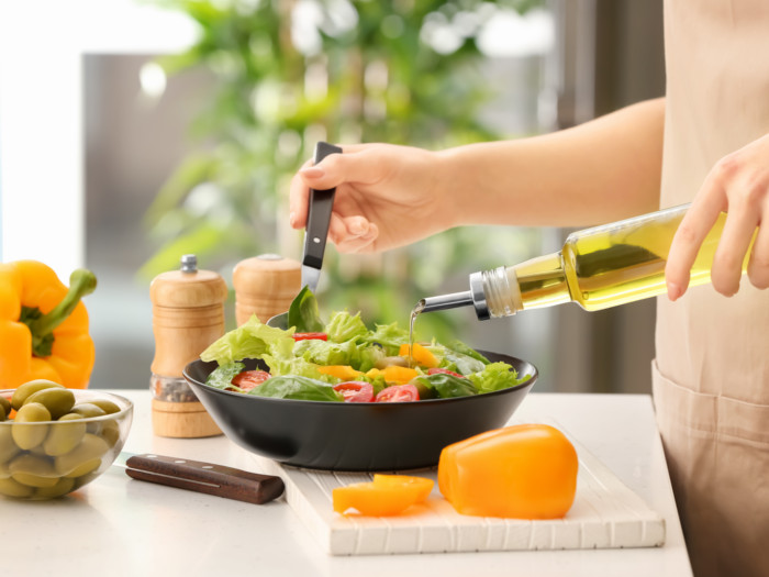 A woman drizzling olive oil on a green salad in a bowl kept on a white kitchen counter