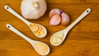 One whole onion, three garlic pods, two spoons containing sliced garlic and garlic powder on a table