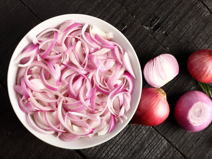 Sliced onions in a bowl, whole onion, and peeled onions on a dark counter