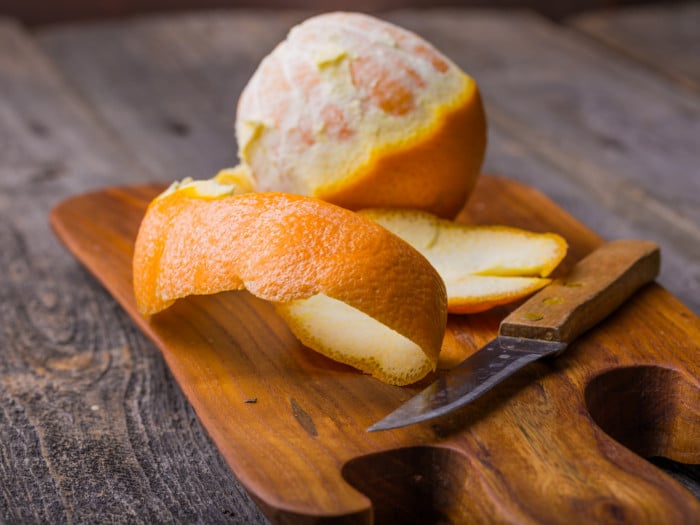 An orange peel, a knife and a partially peeled orange on a wooden board on a wooden surface