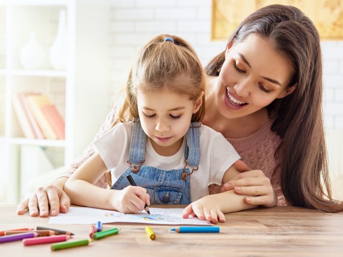 A woman sitting and smiling behind a girl while the girl colors in her coloring book