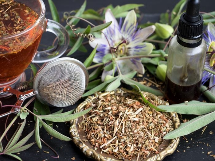 A cup of passion flower tea with a strainer, a container of passion flower oil, and a plate of passion flower powder with fresh flower and leaves on a wooden table