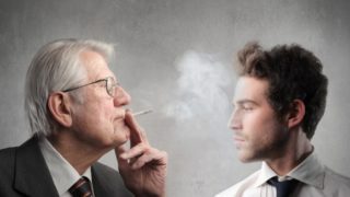 A man smoking with another man looking on.