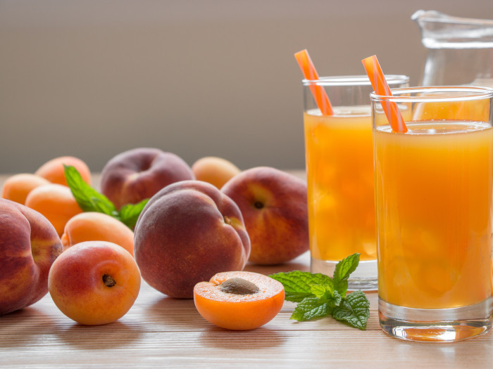 Two glasses of peach juice, placed next to whole peaches with a jug at the back.