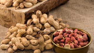 Unshelled peanuts on a brown log and cloth alongside a brass bowl of shelled, raw peanuts