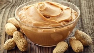 A transparent bowl filled with peanut butter surrounded by raw dried peanuts on a wooden table