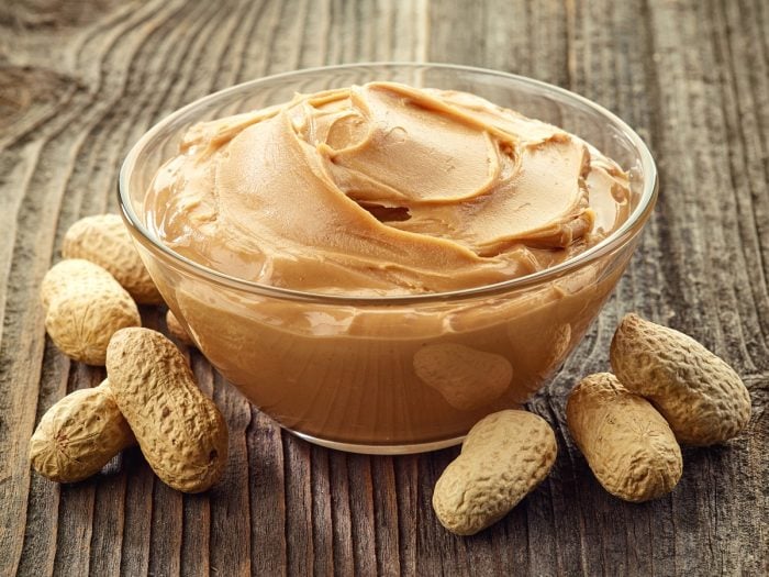 A transparent bowl filled with peanut butter surrounded by raw dried peanuts on a wooden table