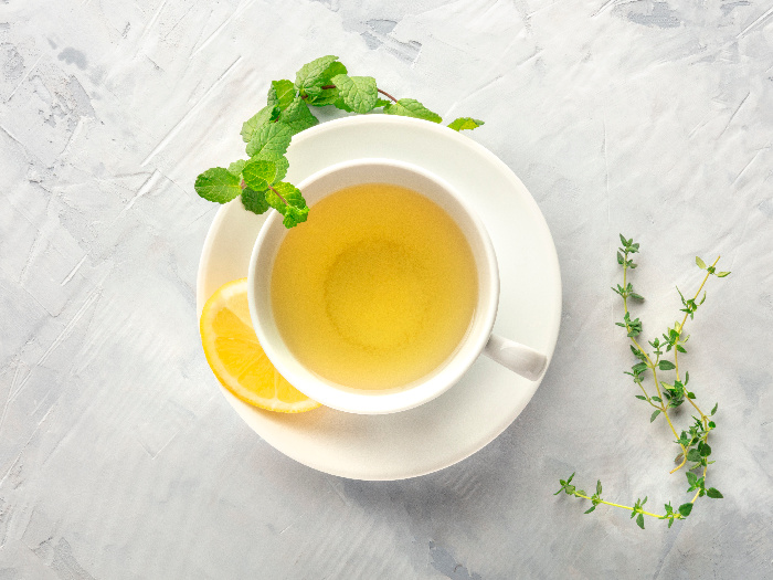 Flatline view of a cup and saucer of tea with a slice of lime, springs of oregano and mint placed on a white surface